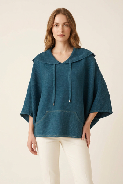 Western model in a three-quarter pose wearing a deep teal pleated cape top and ivory trousers